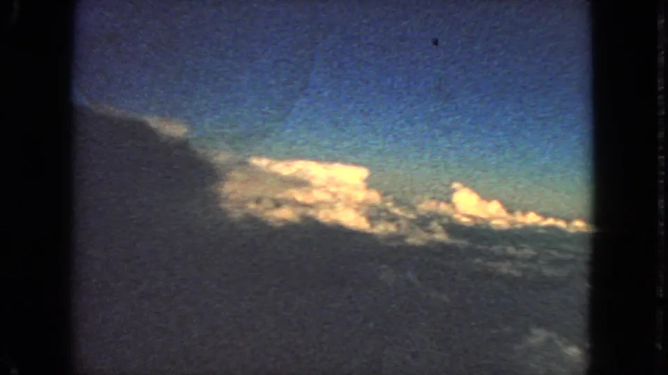 1963 8mm passenger view of blue sky, sunlit clouds, and a dark aircraft window border.