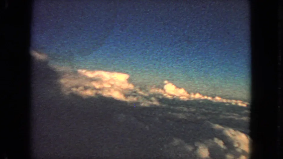 Vintage 8mm aerial view of illuminated clouds beneath blue sky, with dark window framing at the sides.