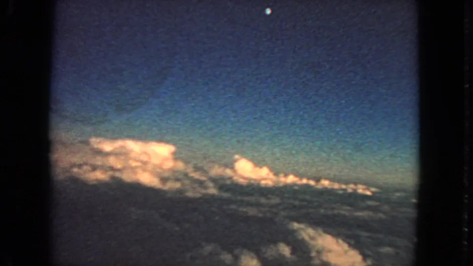 Vintage airplane-window cloud view on 8mm film, with blue sky, sunlit clouds, and visible film texture.