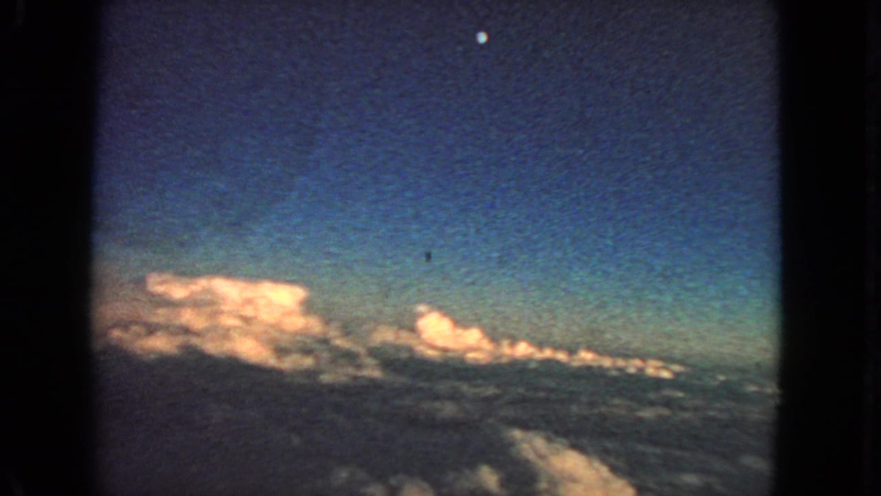 Cloud tops and dark blue sky seen through an airplane-window frame, with a small moon above.