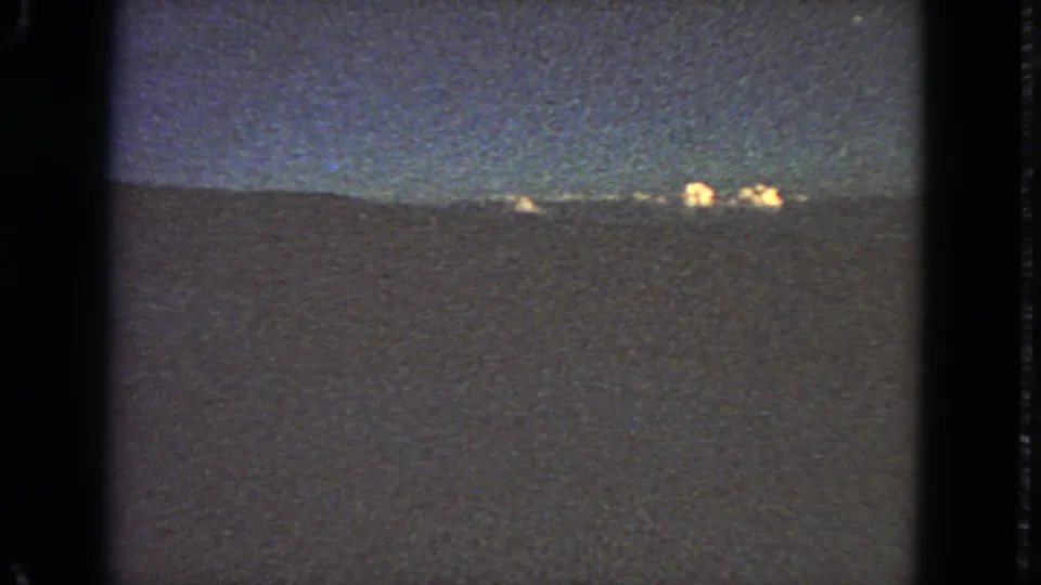 Low-light 8mm frame showing a distant nighttime horizon, scattered warm lights, and strong grain in the sky and foreground.