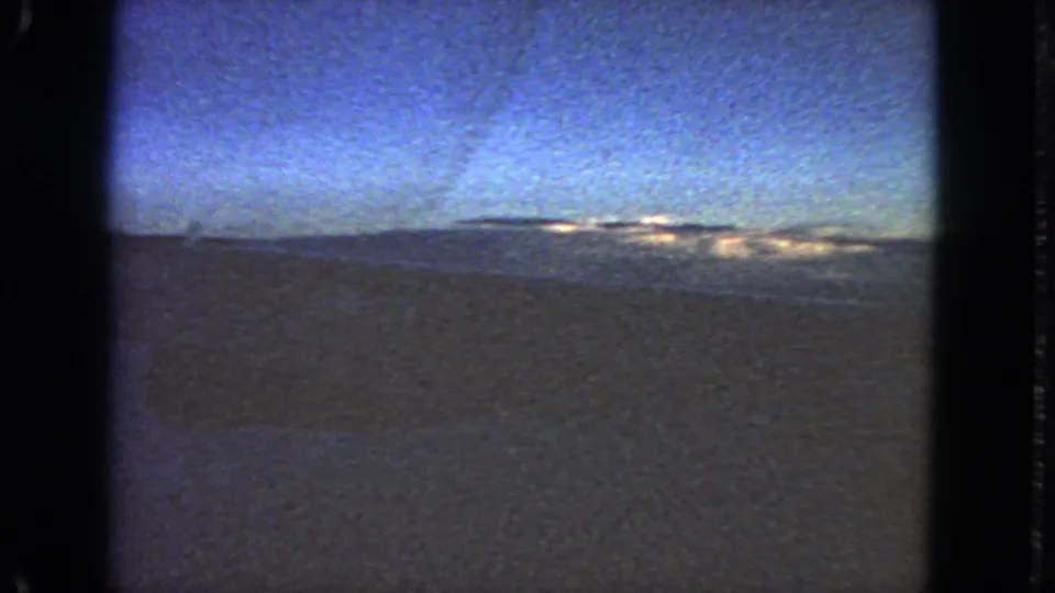 Grainy 8mm night frame with a dark open foreground, blue sky, and small distant lights consistent with the clip metadata location.