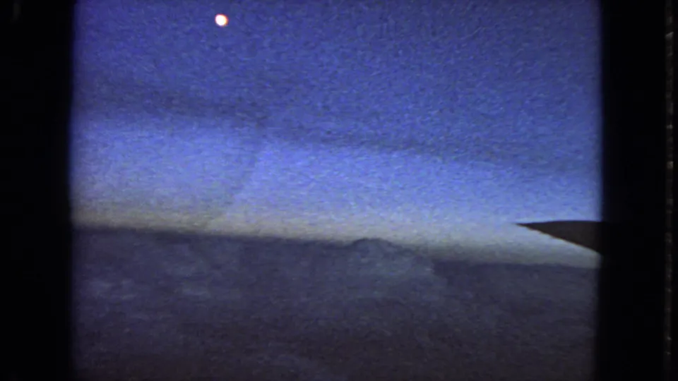 1963 8mm passenger-window view near San Francisco showing grainy blue sky, cloud tops, a small bright point, and wing edge.