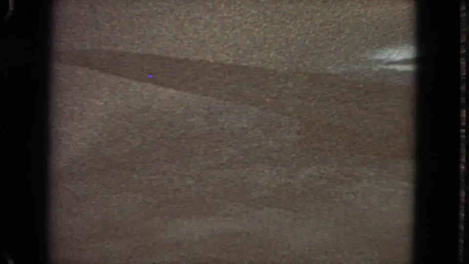 Ambiguous 1963 8mm frame with grainy surface texture, diagonal shadowing, dark borders, and small blue specks in the scan.