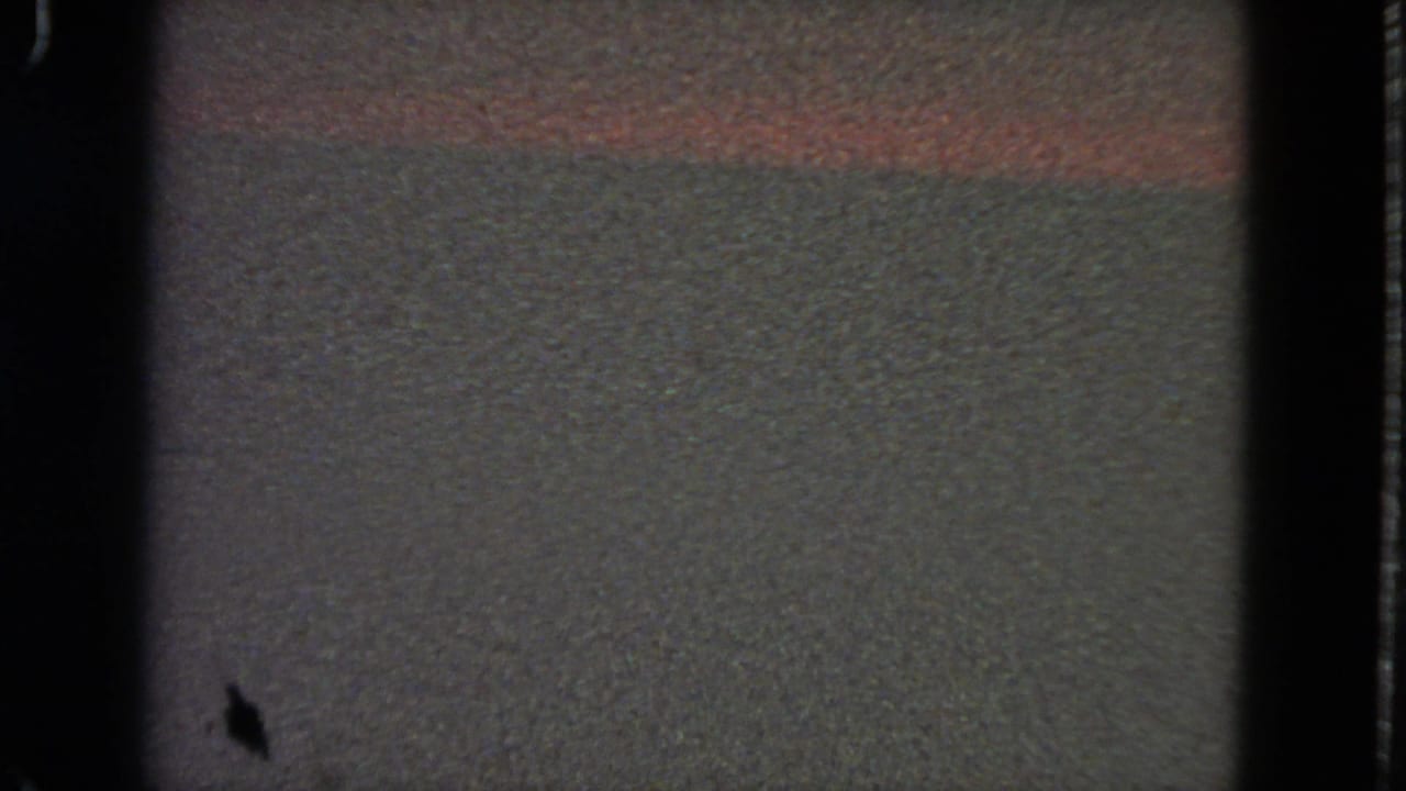 Mostly dark film frame with a small visible portion of muted wall or surface.