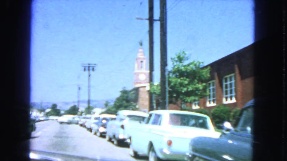 1963 San Francisco street view with parked early-1960s cars, utility poles, a brick building, and a clock tower in the distance.