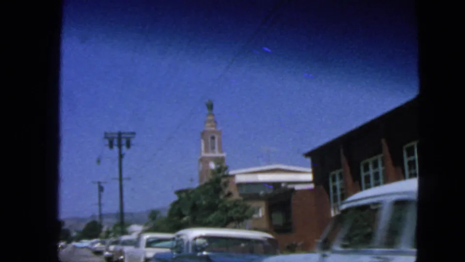 1963 San Francisco view of parked cars beside a brick building, with a clock tower and utility poles framed by 8mm film edges.