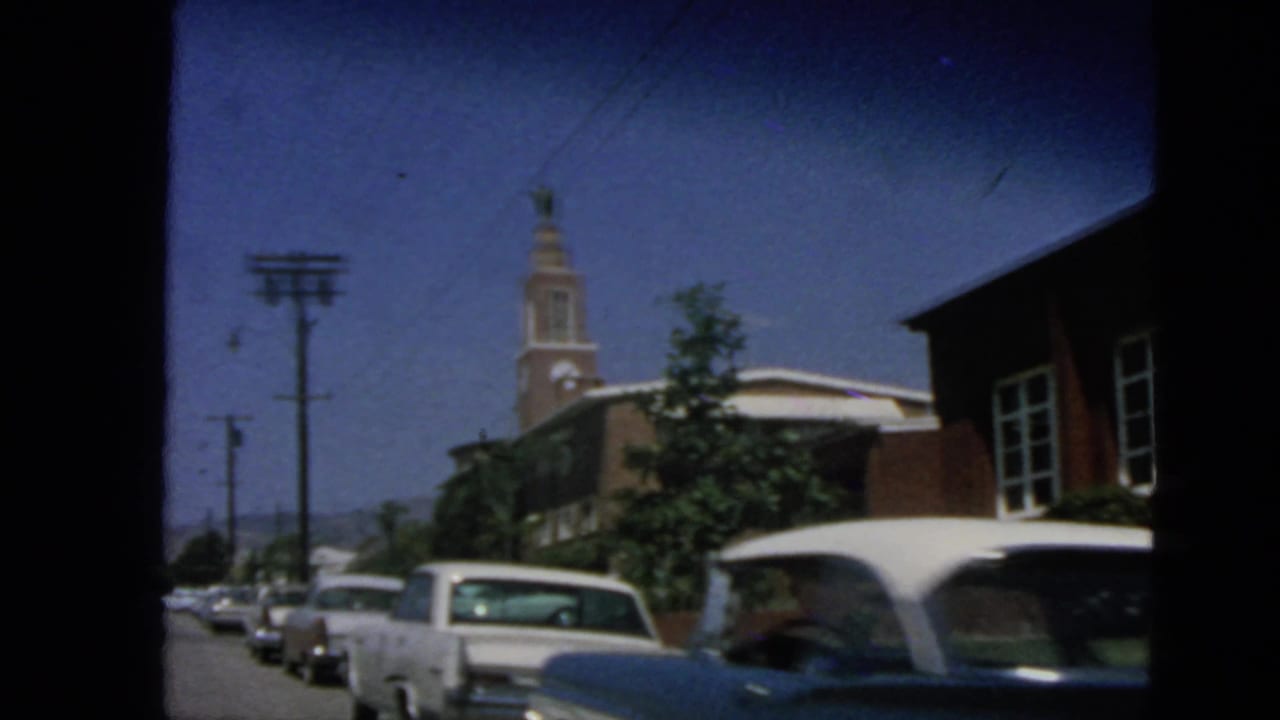 Parked cars line a sunlit street beside a brick building with a clock tower.