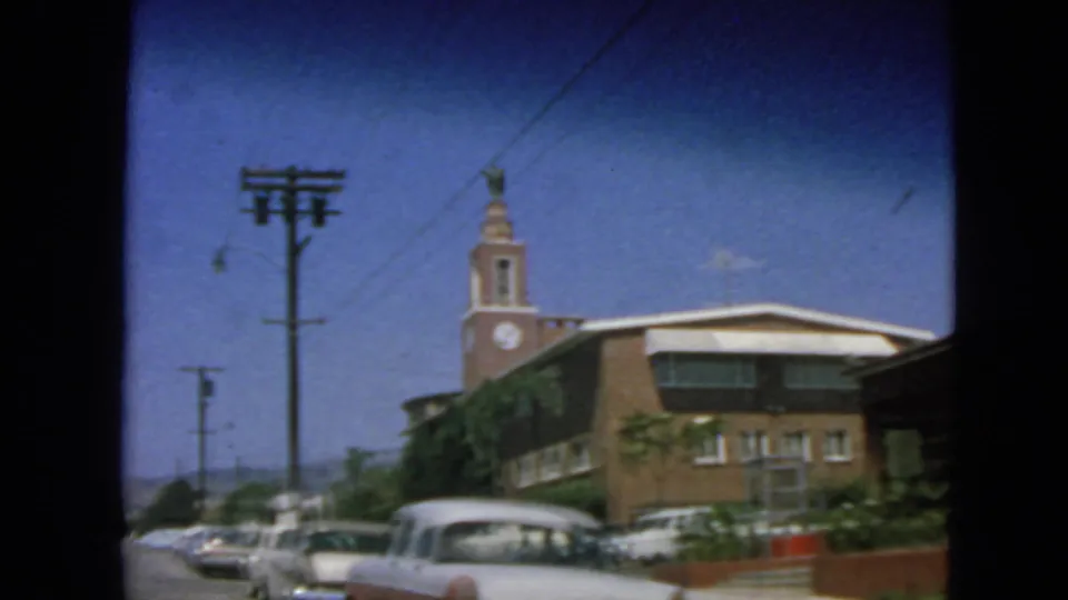 1963 San Francisco exterior showing a brick clock-tower building, parked cars, utility poles, and leafy street plantings.