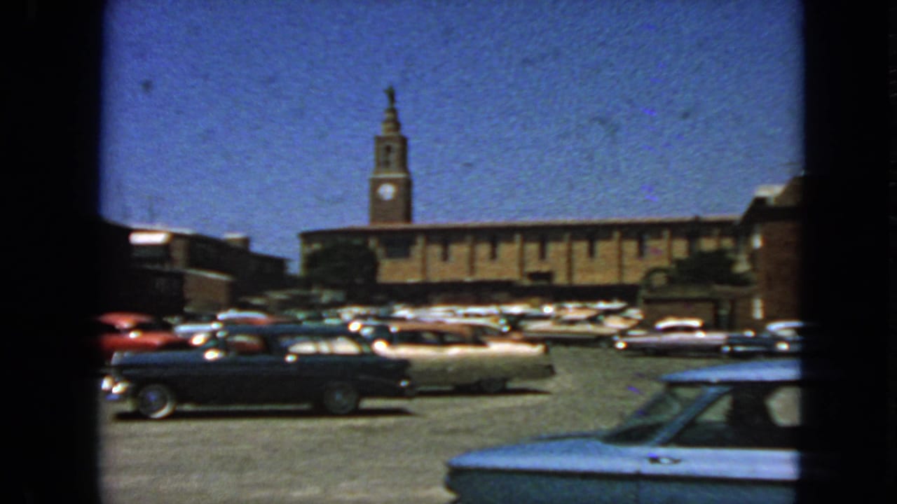 1963 San Francisco home-movie frame showing parked classic cars, a paved lot, and a brick building with a clock tower under blue sky.
