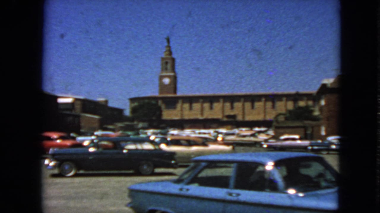 A blue car passes across the foreground of a parking lot filled with older automobiles.