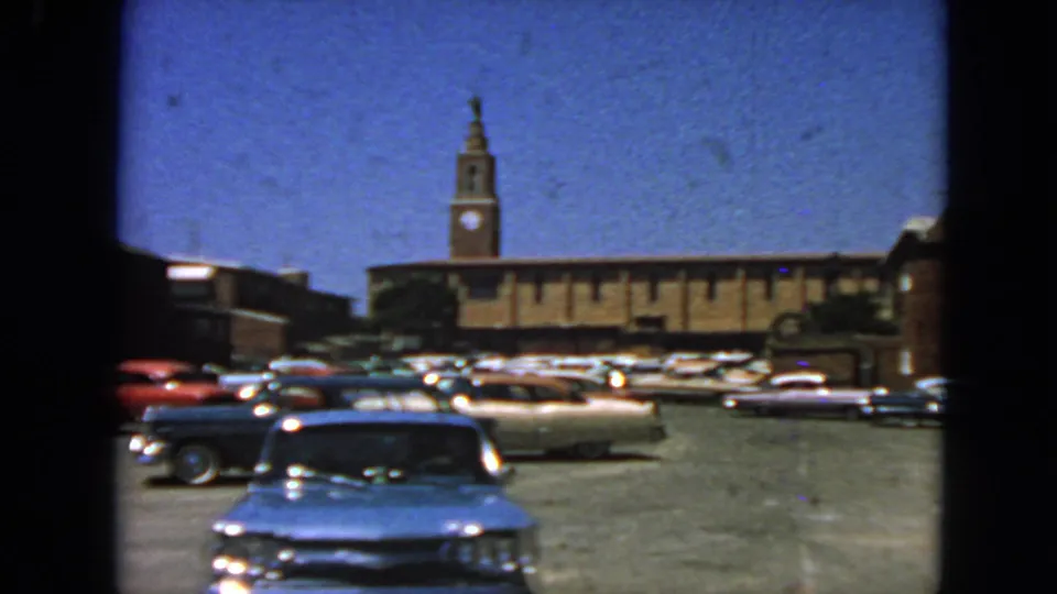 1963 San Francisco home-movie frame showing the front of a blue classic car in a lot with parked cars and a clock tower beyond.