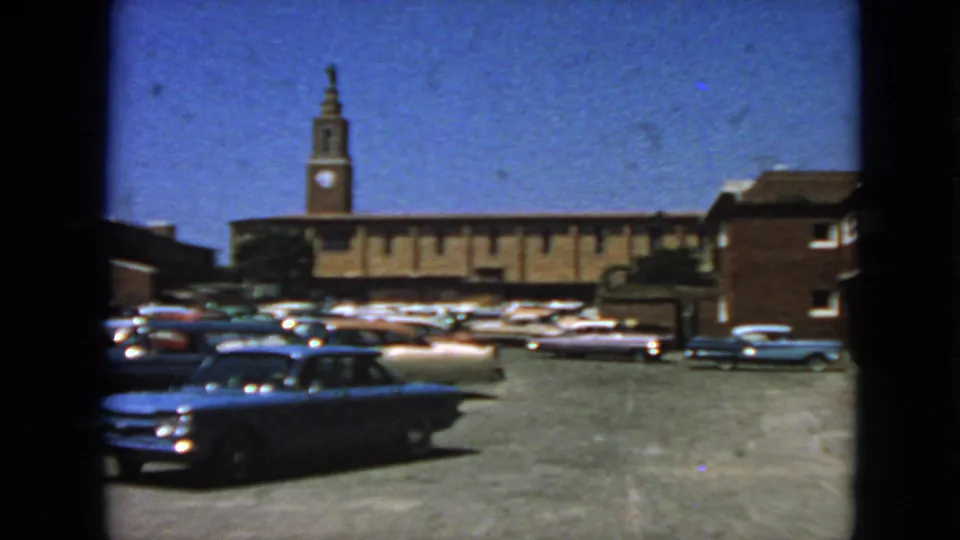 1963 San Francisco frame with a blue classic car moving through a paved lot, with parked cars and a brick clock-tower building behind.