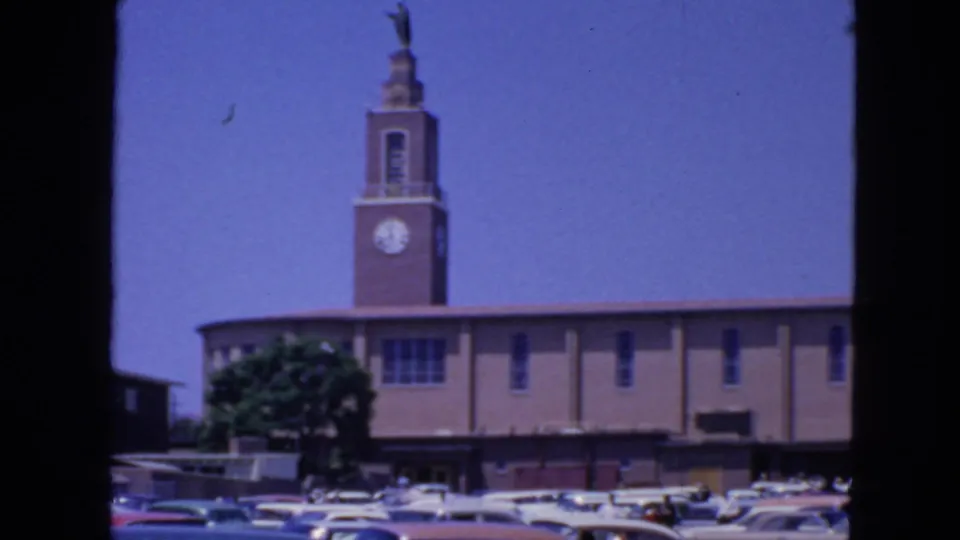 1963 San Francisco home-movie view of a brick building with clock tower, tree, and period cars in front.