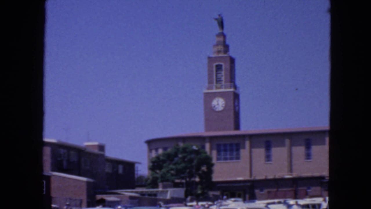 Clock tower with religious statue above a low building under blue sky.
