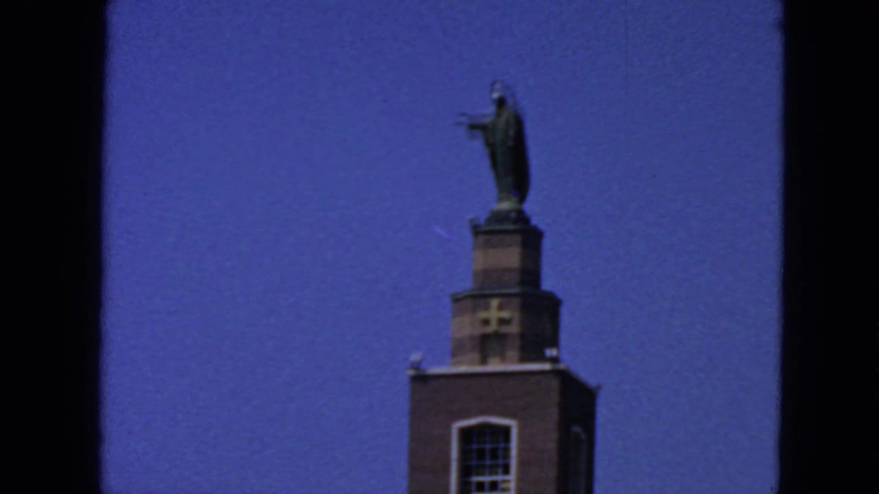 1963 San Francisco home-movie frame showing a brick church tower with cross and robed statue under blue sky.