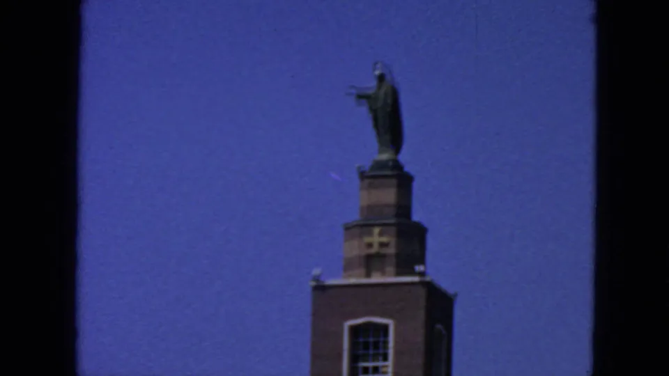 1963 San Francisco home-movie frame showing a brick church tower with cross and robed statue under blue sky.