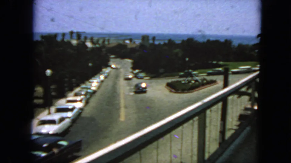1963 San Francisco street view from above, with period cars, a roadside railing, trees, and distant water.