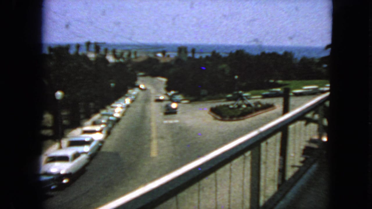 1963 San Francisco home-movie frame from a balcony, showing early-1960s cars along a sunlit street with trees and a distant bay view.