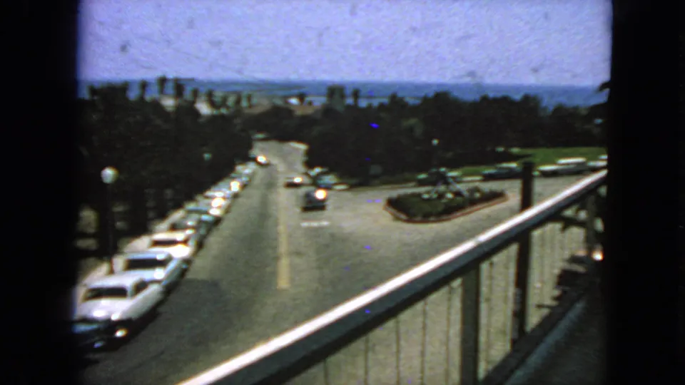 1963 San Francisco home-movie frame from a balcony, showing early-1960s cars along a sunlit street with trees and a distant bay view.