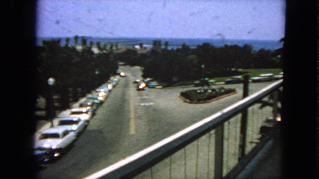 Sunlit intersection seen from above, with parked cars, a few moving cars, trees, and distant water.
