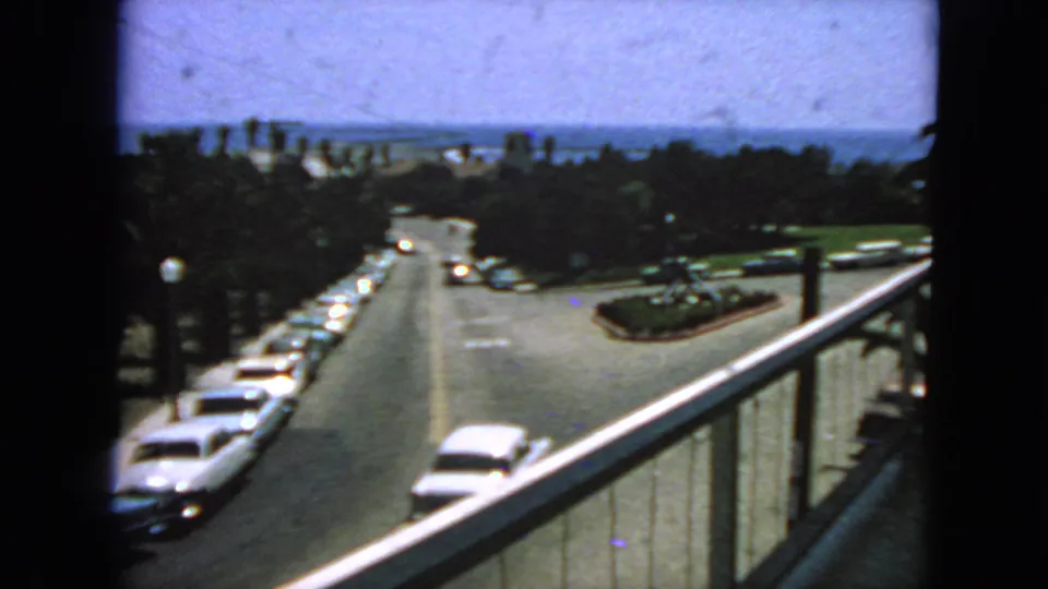 1963 San Francisco balcony view with a white car entering the foreground, parked early-1960s cars, trees, and distant bay scenery.