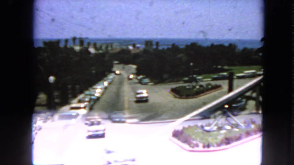 1963 San Francisco home-movie frame showing classic cars on a bright coastal road, with ocean and greenery in the distance.
