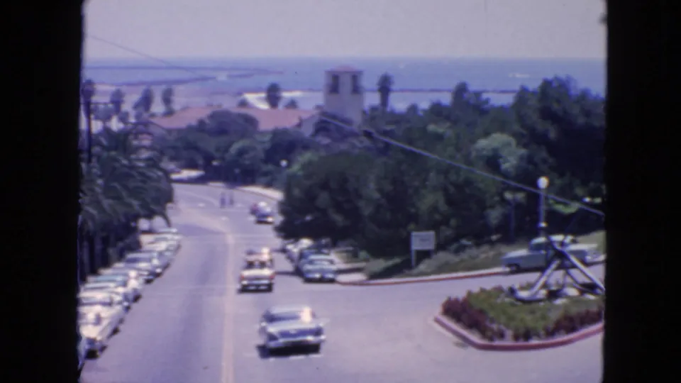 1963 San Francisco street view with classic cars, palm trees, ocean water and a bell-tower-like structure in the distance.