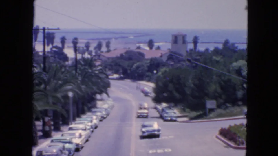1963 San Francisco coastal street scene with parked cars, moving traffic, palms and ocean visible beyond rooftops.
