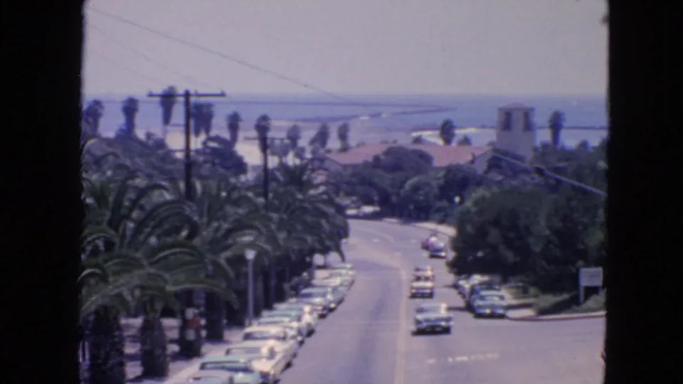 1963 San Francisco home-movie frame showing classic cars on a palm-lined coastal road with ocean views in the distance.