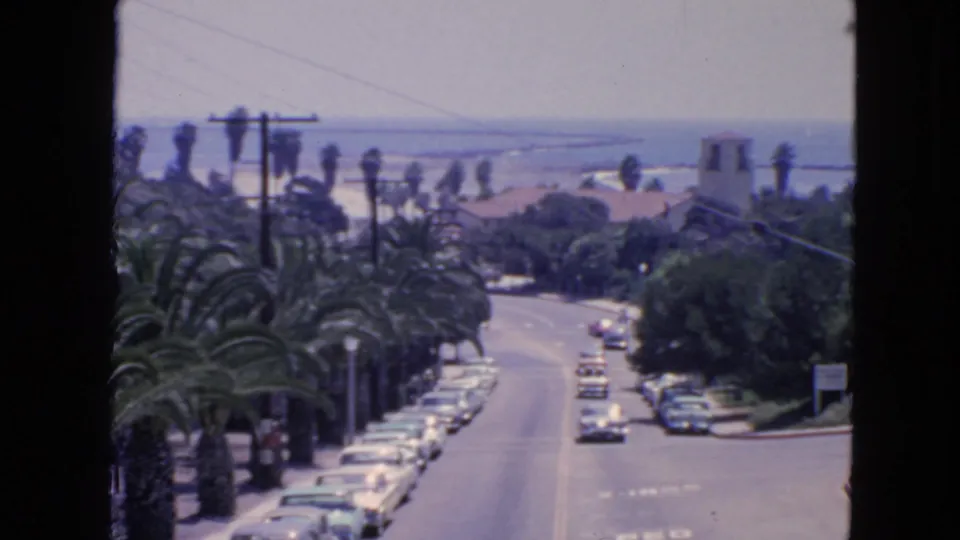1963 San Francisco 8mm view of a sunny palm-lined street with parked cars, moving traffic, rooftops, and water in the distance.