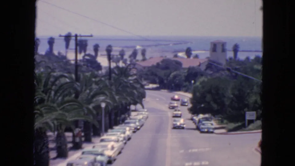 1963 San Francisco street view showing palm trees, parked cars, cars driving uphill or downhill, rooftops, and a hazy waterfront beyond.