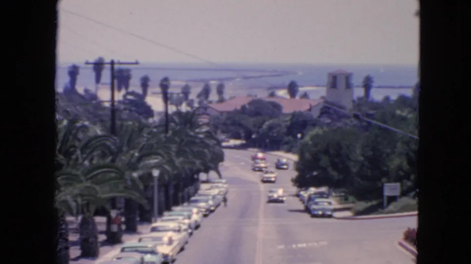 1963 San Francisco 8mm frame of a palm-lined roadway with rows of parked cars, moving vehicles, overhead lines, and water in the background.
