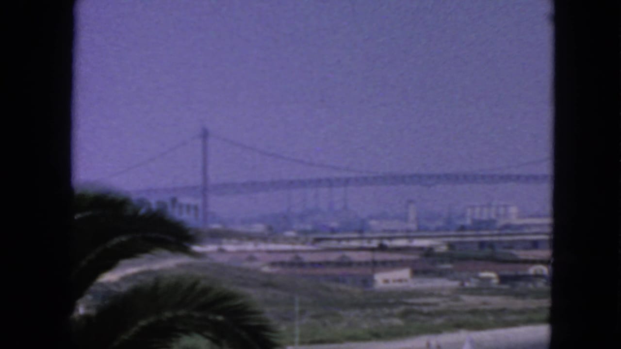 1963 San Francisco home-movie view of a suspension bridge through dark foreground framing, with palm fronds and bayfront industry below.