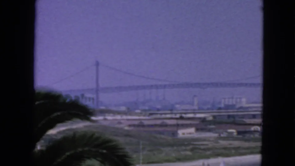 1963 San Francisco home-movie view of a suspension bridge through dark foreground framing, with palm fronds and bayfront industry below.