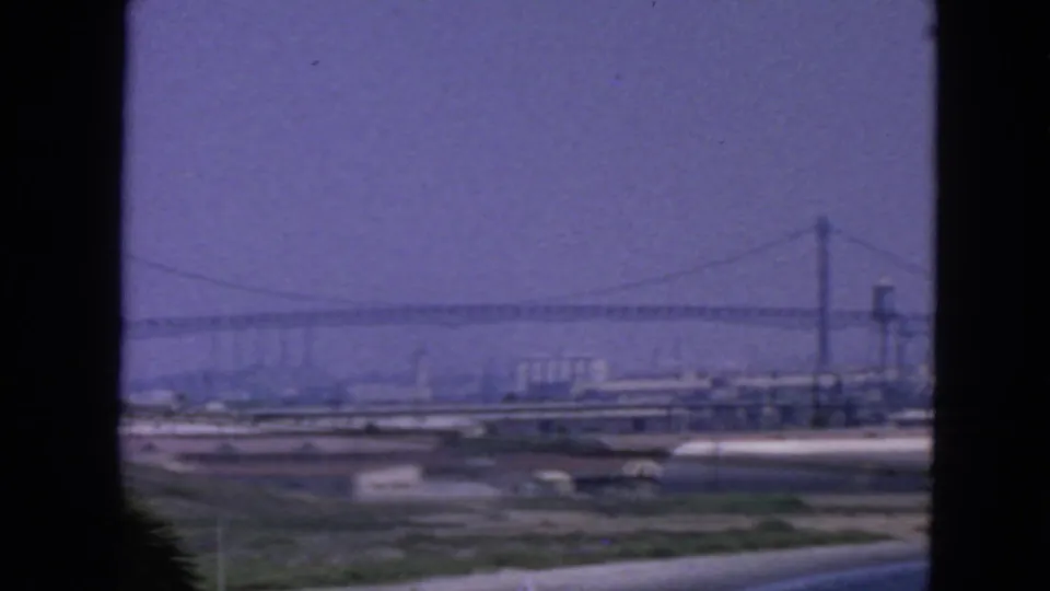 1963 8mm view of a San Francisco bridge tower and span over industrial waterfront lots, seen through dark foreground framing.