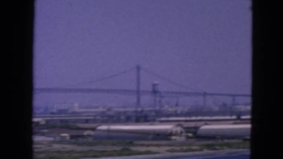 Hazy 1963 San Francisco bridge view with a water tower, low industrial buildings, and dark foreground framing in 8mm color.