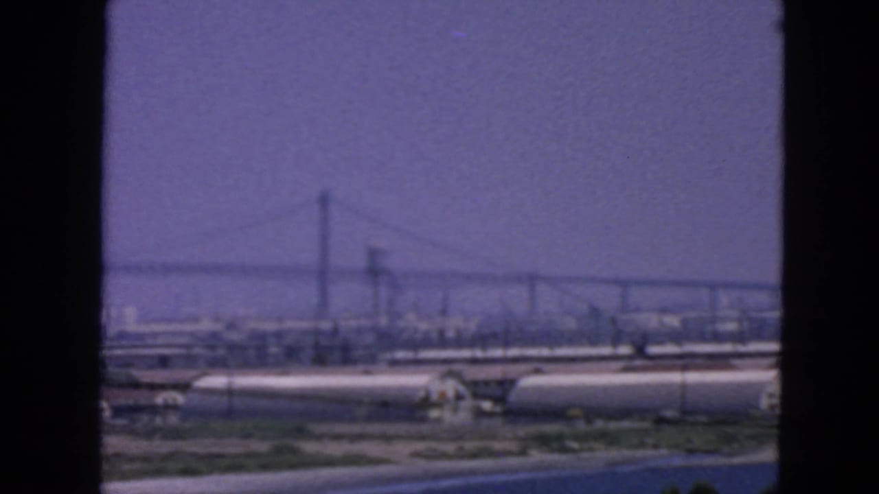 Distant suspension bridge and industrial waterfront tanks under a hazy sky.