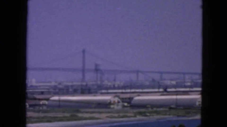 1963 San Francisco waterfront view with a distant suspension bridge, storage tanks, industrial structures, and hazy purple-blue sky.