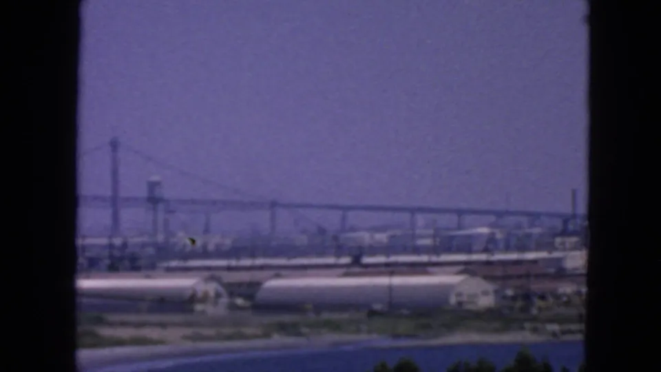 San Francisco Bay industrial waterfront in 1963, with bridge towers and cylindrical storage tanks seen through soft 8mm focus.