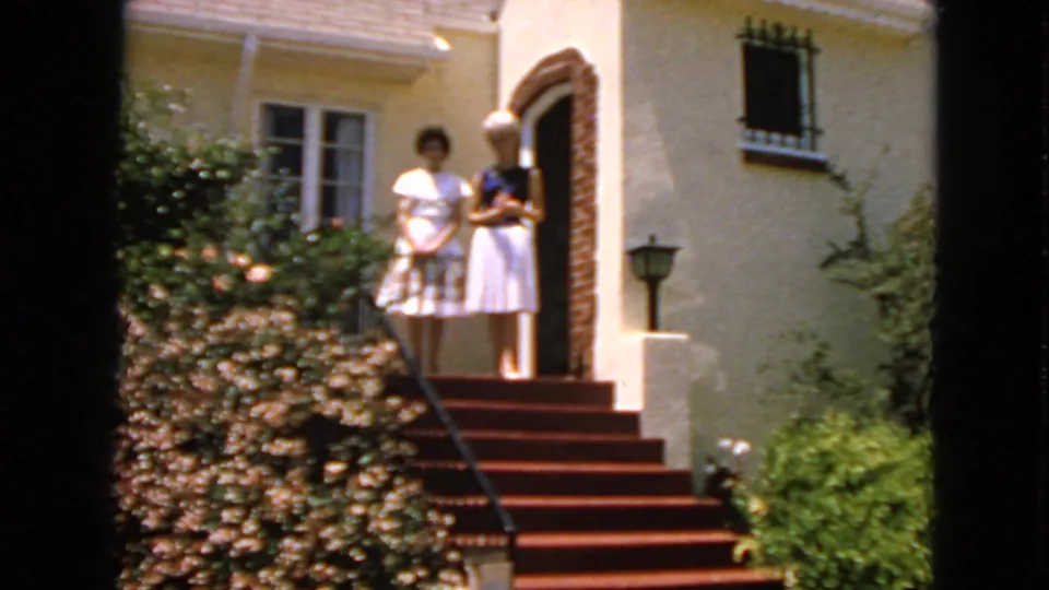 Two women in light dresses pose on the front steps of a stucco California house, with shrubs and a brick entry arch visible.