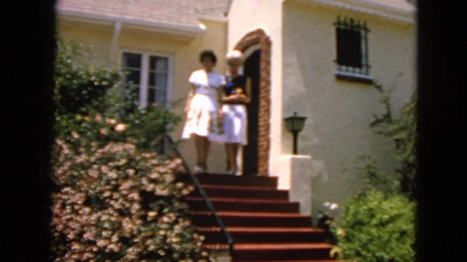 A 1963 home-movie frame shows two women standing together on red house steps beside a brick arch and barred window.