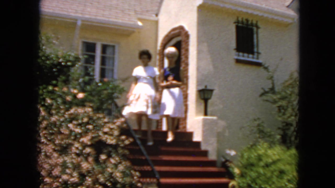 Two young women stand on red-carpeted steps outside a cream-colored house.