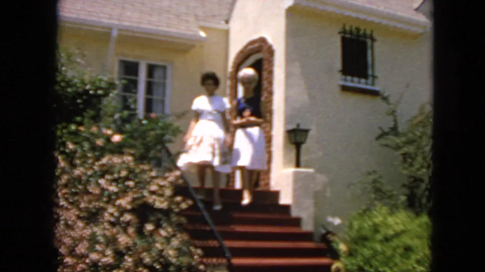 Two young women in 1960s-style dresses stand on the front steps of a San Francisco residence, with shrubs and an arched doorway nearby.