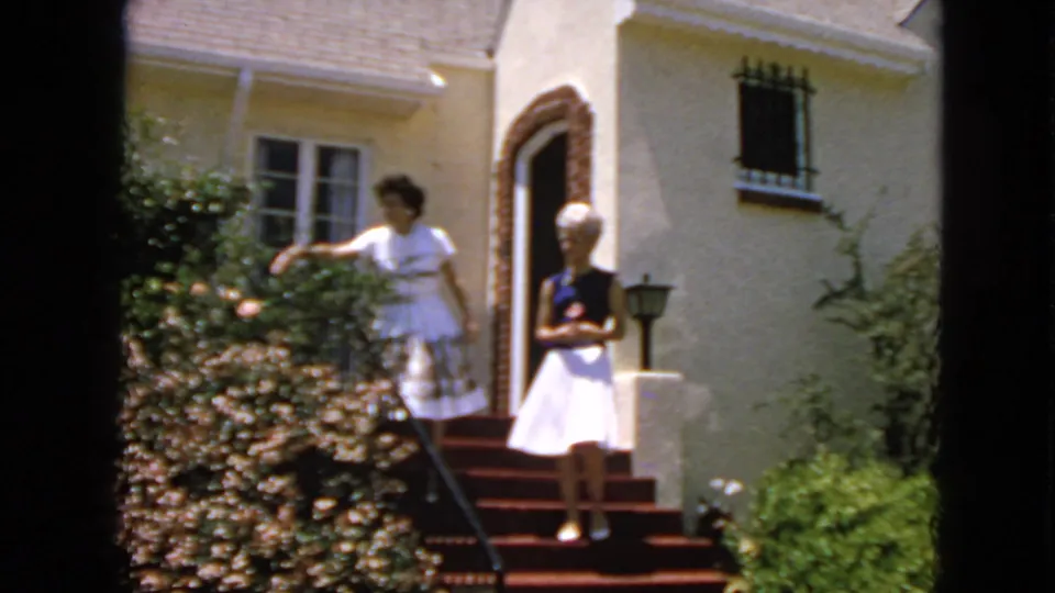 Outside a San Francisco home, one young woman reaches toward flowers while another waits on the red-carpeted steps near the open doorway.