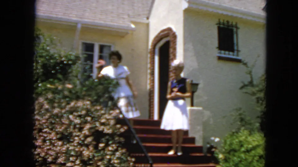A young woman in a white dress stands beside a flowering shrub at a San Francisco residence while another woman stands on the front steps.