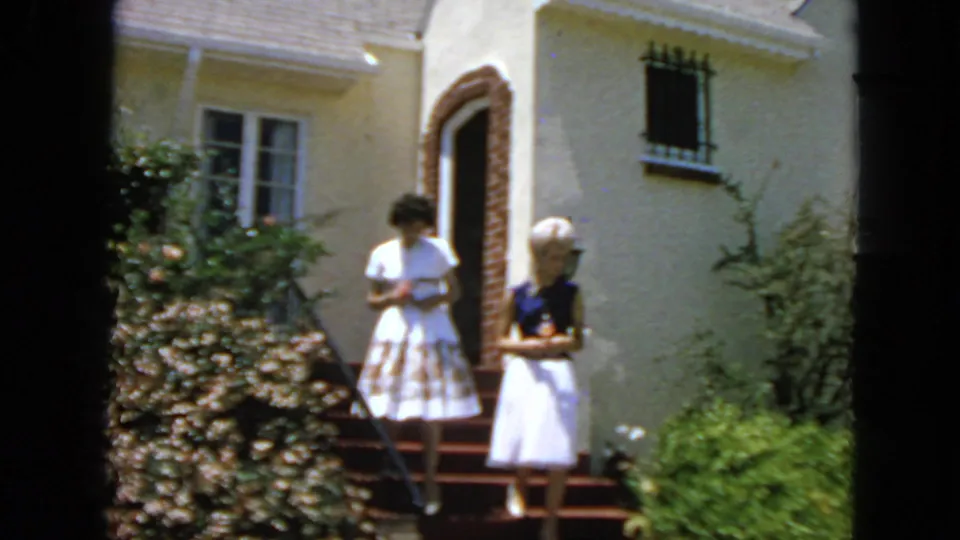 Two young women stand together on the red-carpeted front steps of a San Francisco home, framed by shrubs, stucco walls, and a dark doorway.