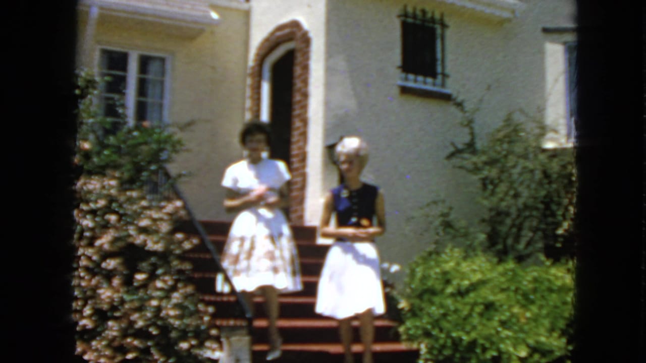 Two adult women in dresses stand by a residential entrance in San Francisco, 1963, with shrubs and red-carpeted steps visible.