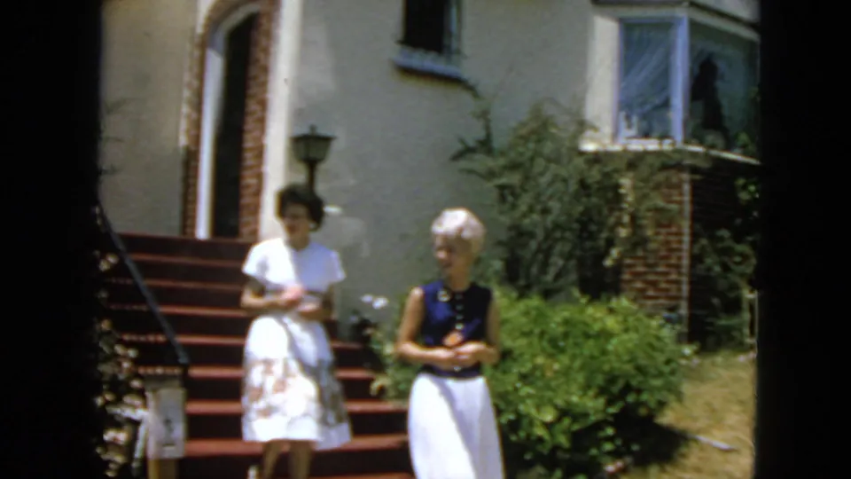 Two adult women descend red-carpeted residential steps in 1963 San Francisco, one in a white dress and one in a navy top with white skirt.