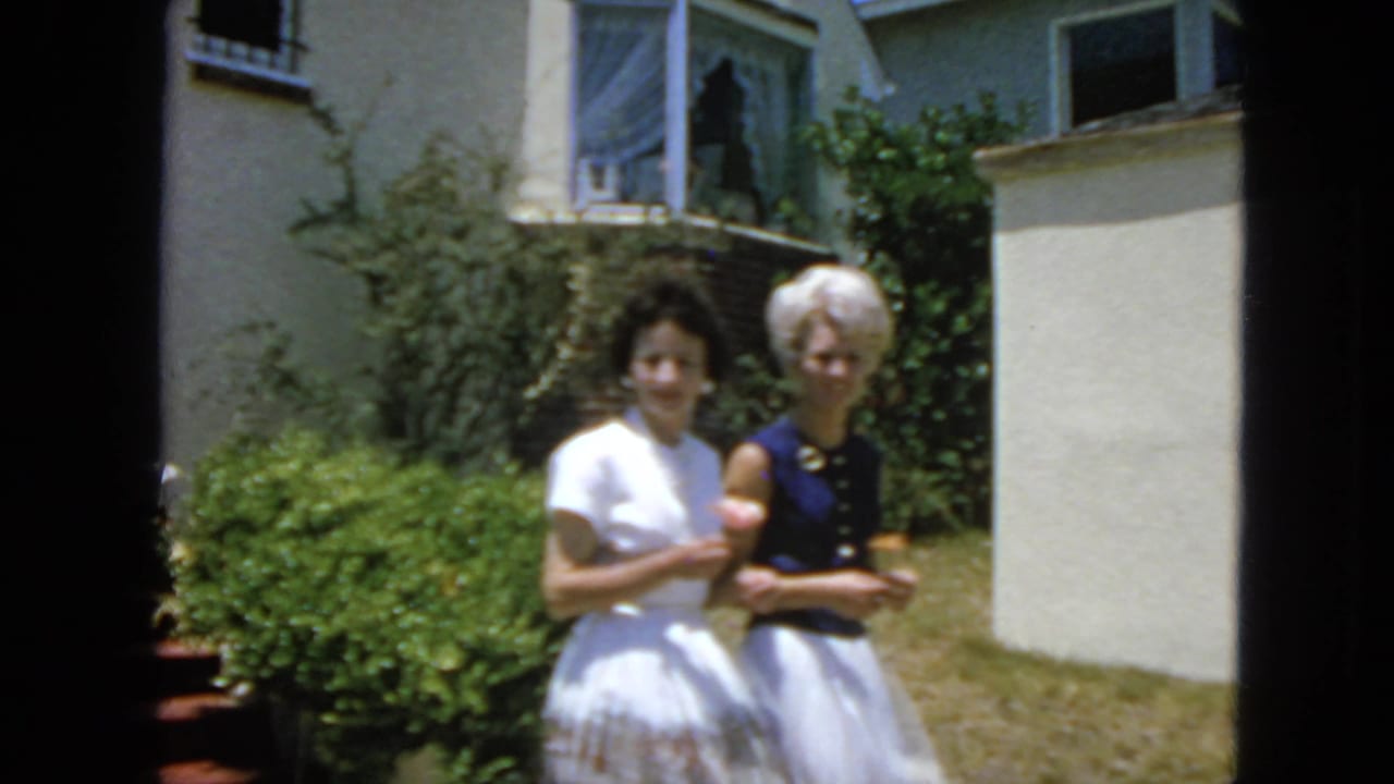 In 1963 San Francisco home-movie footage, two women stand in a sunny residential yard near shrubs and a light-colored house with a small cat.
